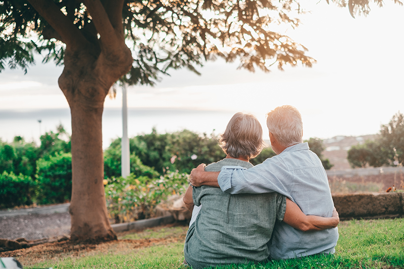 Happy grey haired middle aged woman snuggling to smiling older husband, enjoying tender moment at park. Bonding loving old family couple embracing, looking sunset